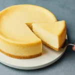 Slice of classic New York-style cheesecake being served from a whole cake on a white plate