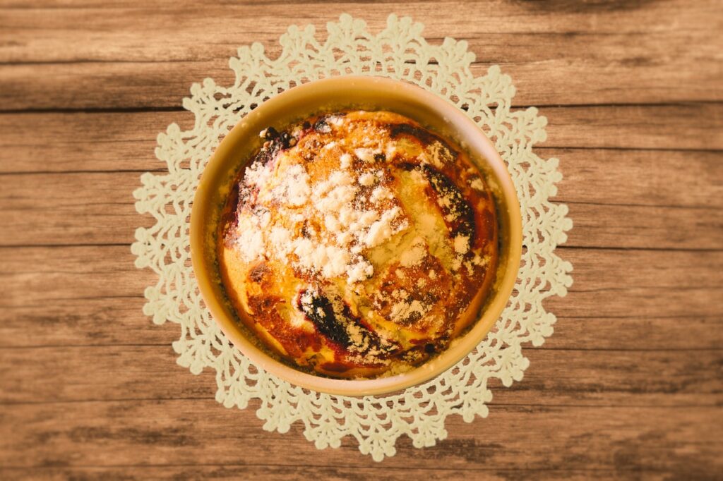 Rustic kitchen setting with ingredients for cobbler topping, including flour, sugar, butter, baking powder, milk, and fresh fruits like peaches and berries, alongside a partially prepared cobbler in a cast-iron skillet.